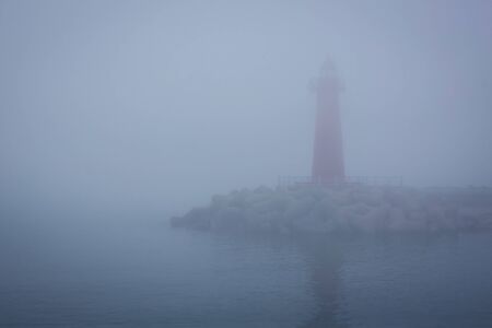 Thick fog, early morning. Unclear outline of the lighthouse. Around the water, the sea. Blue tinting.の写真素材