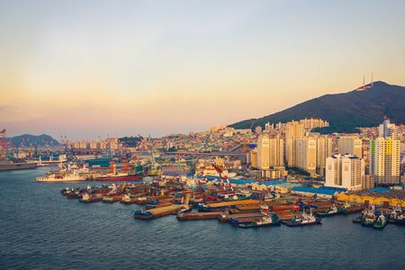 Busan /  South Korea - 07.06.2019: Busan panorama, view at sunset. There are many ships in the harbor. On the shore there are tall buildings, in the distance a hill, a mountain. View from the water.のeditorial素材