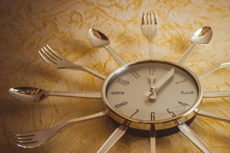 Kitchen, a metal clock on the wall, the arrows show noon. Meals, lunch, diet. Light golden background, bottom view, vignetting. The product of mass production.の写真素材