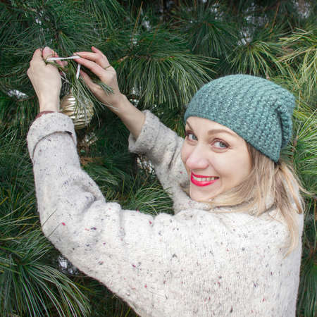 Waiting for the holiday. Decorating a Christmas tree in the yard. A woman in a hat hangs a golden ball on green branches.の写真素材