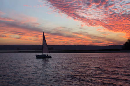 Beautiful dramatic sunset over the river. Sailboat on dark water. In the distance the shore. Blue sky, red and purple clouds.の写真素材
