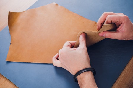 Working day in studio. Man leather worker drawing sketch of purse in notebook on table with leather wallets and tools for sewing. Effective work in leather workshop. Design of fashion leather purseの写真素材