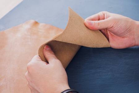 Working day in studio. Man leather worker drawing sketch of purse in notebook on table with leather wallets and tools for sewing. Effective work in leather workshop. Design of fashion leather purseの写真素材