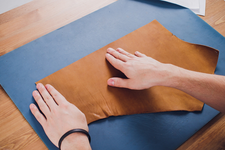 Working day in studio. Man leather worker drawing sketch of purse in notebook on table with leather wallets and tools for sewing. Effective work in leather workshop. Design of fashion leather purseの写真素材