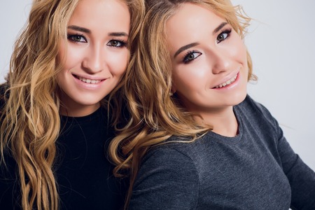 Two beautiful women in black night fashion dress posing isolated on a white gray background. Pretty brunette girl friends twins having fun drinking cocktails. Singing and dancing. Festive mood. Insideの写真素材