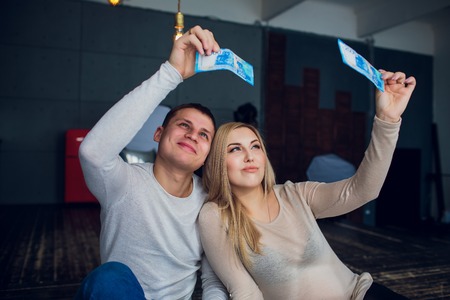 Pregnant family with money and babys clothes sit on a sofa of the house. Family budget.