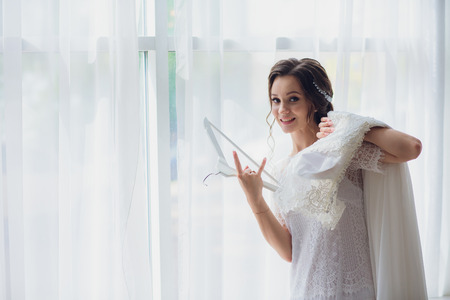 Pretty brunette bride in silk dressing gown and lacy veil holding wedding dress on a hanger near the window.の写真素材