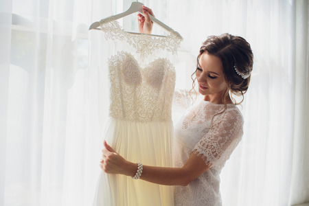 Pretty brunette bride in silk dressing gown and lacy veil holding wedding dress on a hanger near the window.の写真素材
