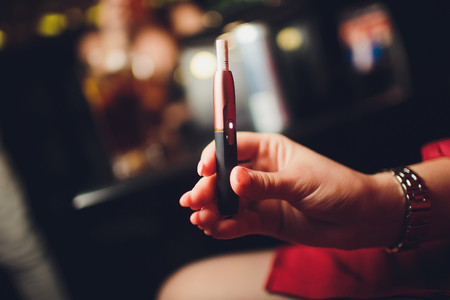 Electronic cigarettes, technology cigarette. Tobacco system IQOS Female hand on blurred background of the restaurant. red case.の写真素材
