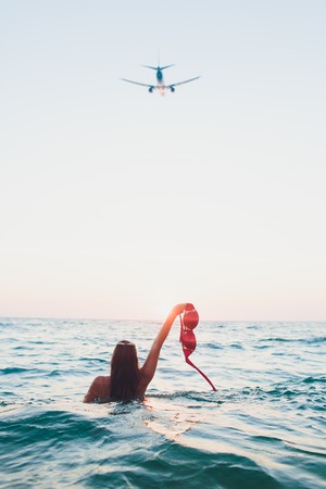 Young woman with long hair, blonde, topless, sitting in the water and holding in his hand a bikini top in the sunshine. took off her bra, plane, seaの写真素材
