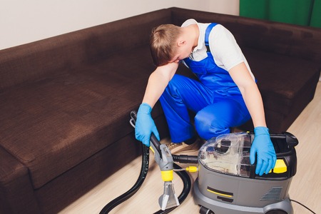 Tired young man sitting on sofa in living room. extractor, couch couch, chemistry, washing, cleaning. working blue uniformの写真素材