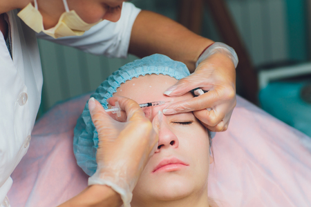Doctor giving face lifting injection on mid age woman in the forehead between eyebrows to remove expression wrinkles in a clinic surgery room O.R background Beautify filler for face anti aging concept.の写真素材