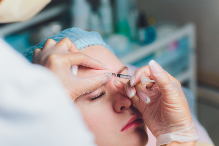 Doctor giving face lifting injection on mid age woman in the forehead between eyebrows to remove expression wrinkles in a clinic surgery room O.R background Beautify filler for face anti aging concept.の写真素材