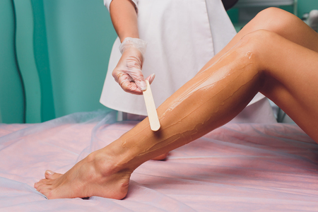 The beautician is preparing for depilation and applying the cream with wax stick on the beautiful female legs. Beauty salonの写真素材