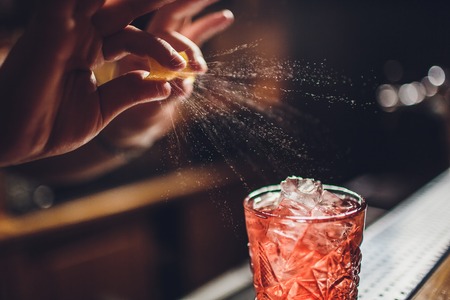 Barman s hands sprinkling the juice into the cocktail glass filled with alcoholic drink on the dark...
