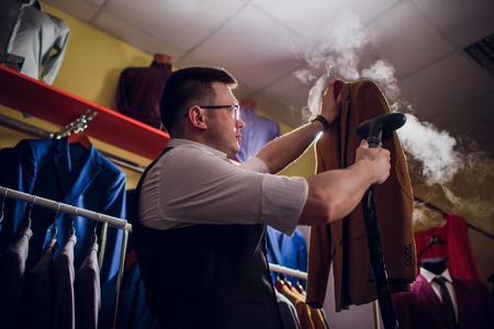 tailor cuts and sews a suit. A young man in the tailoring shop makes mens clothing. Custom-made clothing workshop. holds in her hand an electric iron. wet-heat treatmentの写真素材
