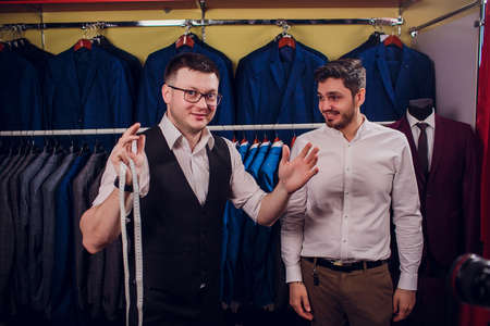 Businessman in classic vest against row suits in shop. Man helps another try on suit in clothing storeの写真素材