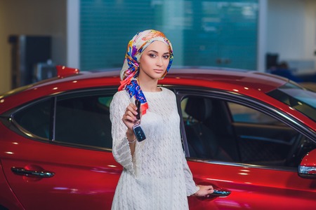 property and people concept - muslim woman in hijab with car key over car show background. happy woman taking car key from dealer in auto show or salonの写真素材