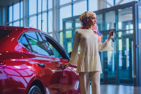 property and people concept - muslim woman in hijab with car key over car show background. happy woman taking car key from dealer in auto show or salonの写真素材