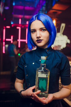 Beautiful cheerful brunette bartender girl in white shirt and black bow tie, serving alcohol drink at nightclub bar, holding bottle in hand, pouring drink in glassの写真素材