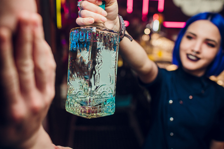 Beautiful cheerful brunette bartender girl in white shirt and black bow tie, serving alcohol drink at nightclub bar, holding bottle in hand, pouring drink in glassの写真素材