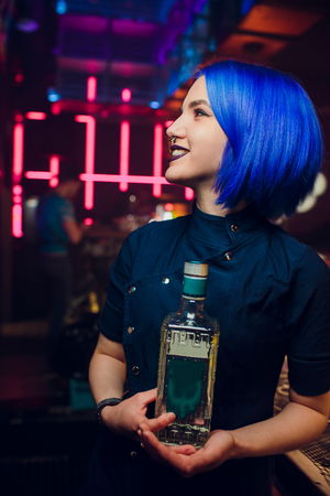 Beautiful cheerful brunette bartender girl in white shirt and black bow tie, serving alcohol drink at nightclub bar, holding bottle in hand, pouring drink in glassの写真素材