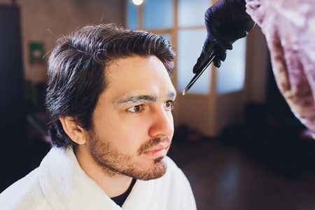 Closeup of process of threading procedure in barber shop. Professional barber correcting shape of brows with threads to smiling young man sitting in chair. Concept of eyebrows care.の写真素材