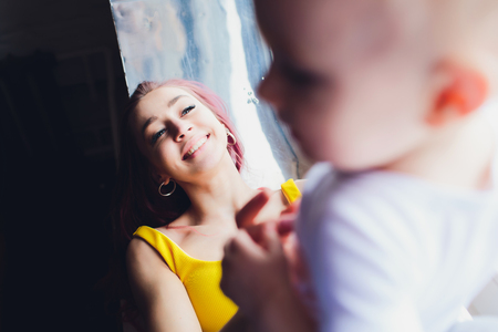 Loving mother playing with her baby sitting on a window.の写真素材