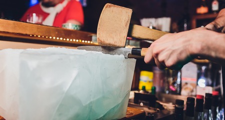 Bartender is cutting a large block of ice to prepare whiskey cocktails. Selective focus on the ice block. Hospitality and fine whiskey conceptの写真素材