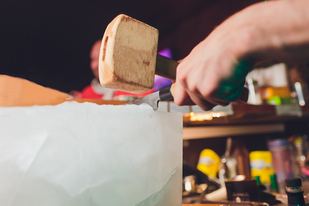 Bartender is cutting a large block of ice to prepare whiskey cocktails. Selective focus on the ice...