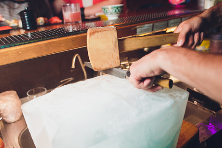 Bartender is cutting a large block of ice to prepare whiskey cocktails. Selective focus on the ice block. Hospitality and fine whiskey conceptの写真素材