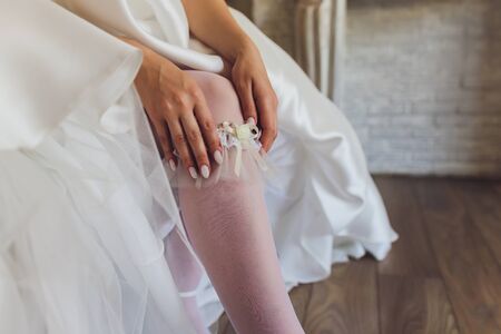 In the morning, the bride in stockings and a white wedding dress wears a garter on her leg, the bride is holding her hands for the garter.の写真素材