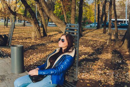 A beautiful girl is sitting on a park bench on a background of green nature.の写真素材