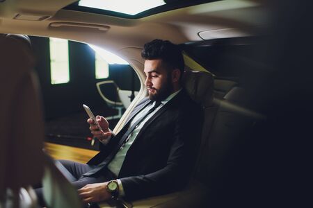 Busy working day. Handsome young man in full suit looking at his smart phone while sitting in the...
