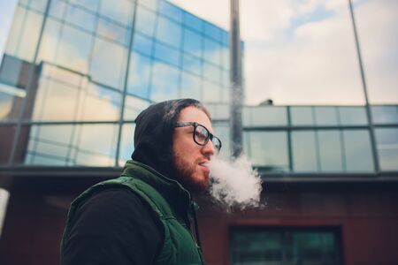 Portrait of young guy with large beard in glasses vaping an electronic cigarette opposite urban background.の写真素材