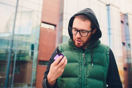 Portrait of young guy with large beard in glasses vaping an electronic cigarette opposite urban...