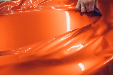 Car wrapping specialist putting vinyl foil or film car wrapping protective film yacht, boat, ship, car, mobile home. orange film heating with hair dryer and trimming plastic soft hard squeegeeの写真素材