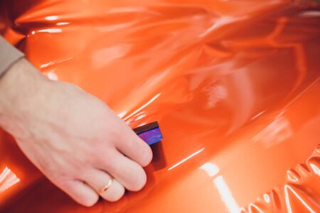 Car wrapping specialist putting vinyl foil or film car wrapping protective film yacht, boat, ship, car, mobile home. orange film heating with hair dryer and trimming plastic soft hard squeegeeの写真素材