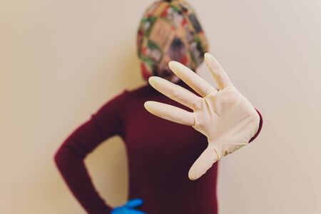 Girl in medical mask and gloves is looking at cameraand show her palm in stop gesture. Concept of fighting against virus due pandemic of coronavirus.の写真素材