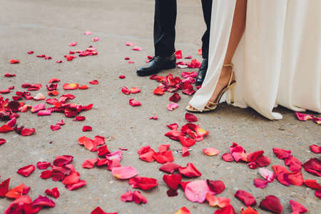 legs and feet of bride and groom with white dress and black trousers, some red and pink rose petals are lying on the ground.の写真素材