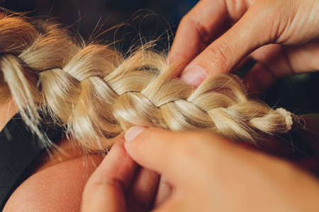 Process of braiding the master weaves braids on her head blond little girl in beauty salon close up. Professional hair care and creating hairstyles.の写真素材