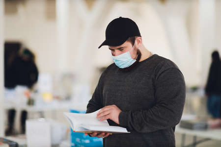 Man learning studying reading book in library during coronavirus covid19 pandemic, wearing protective medical face mask. New normal for librarian.の写真素材