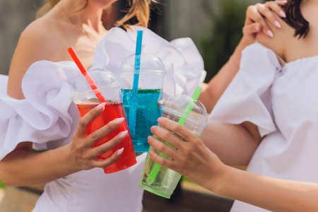 Three ladies cheering with tasty nectars with ice cubes, with black straws, palms decoration, in swim pool, transparent clean clear blue water, sun shines, smooth tanned skin, carefree festive mode.の写真素材