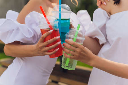 Three ladies cheering with tasty nectars with ice cubes, with black straws, palms decoration, in swim pool, transparent clean clear blue water, sun shines, smooth tanned skin, carefree festive mode.の写真素材