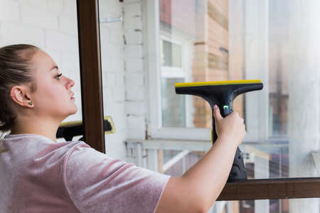 Beautiful young housewife in working clothes washing windows by professional tools at home.の写真素材