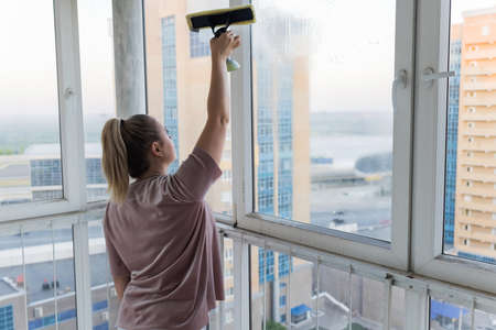 Beautiful young housewife in working clothes washing windows by professional tools at home.の写真素材