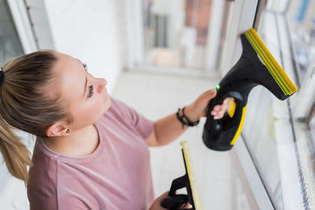 Beautiful young housewife in working clothes washing windows by professional tools at home.の写真素材