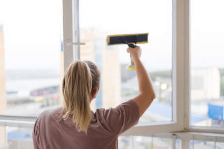 Beautiful young housewife in working clothes washing windows by professional tools at home.の写真素材