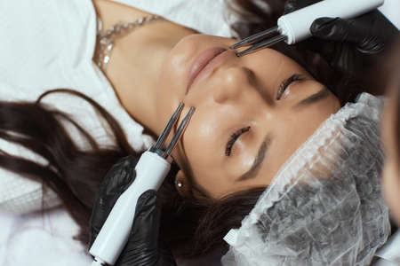 Cosmetology. Beautiful Woman At Spa Clinic Receiving Stimulating Electric Facial Treatment From Therapist.の写真素材