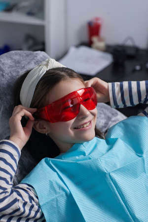 Little girl sits in dental chair and wears protective glasses. She dabbles.